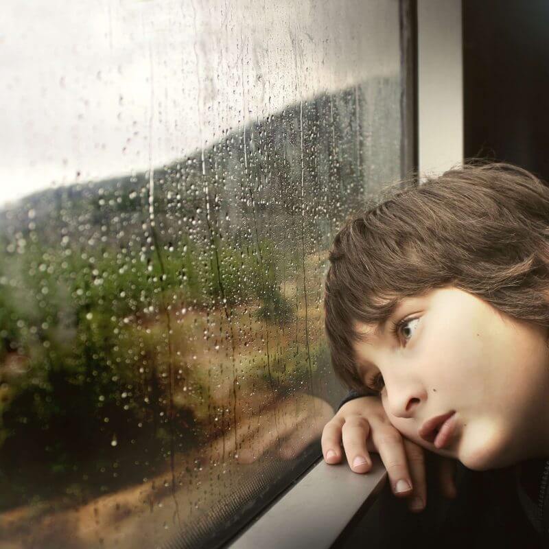 Ein Kind legt den Kopf auf die Fensterbank Draußen regnet es